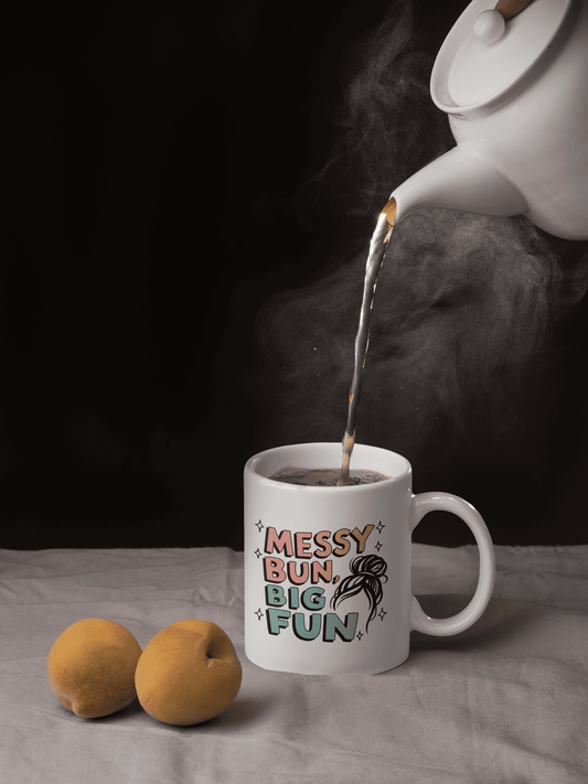White ceramic mug with “Messy Bun, Big Fun” design, being filled with hot tea from a teapot, styled with two peaches on a soft fabric