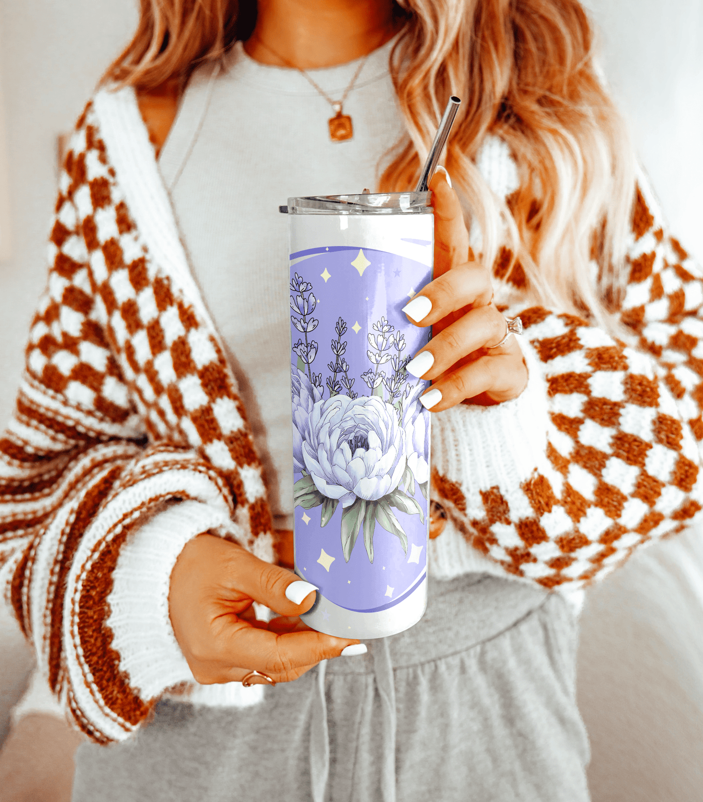 Woman holding a lavender floral tumbler with a metal straw, wearing a cozy checkered cardigan. Only her torso and hands are visible.