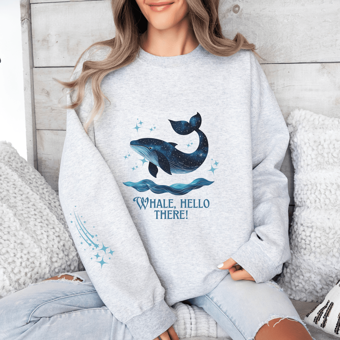 Woman wearing a light grey sweatshirt featuring a galaxy-style whale jumping from ocean waves with the quote “Whale, Hello There!”