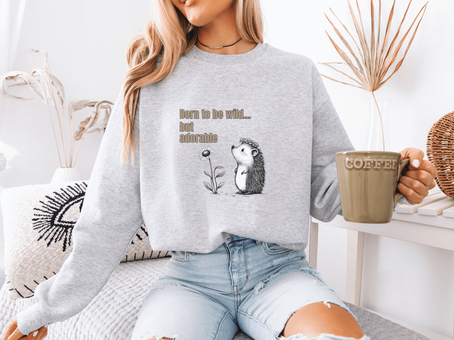 Woman wearing a light gray sweatshirt with “Born to be wild… but adorable” text and a cute hedgehog graphic, sitting on a cozy white couch.