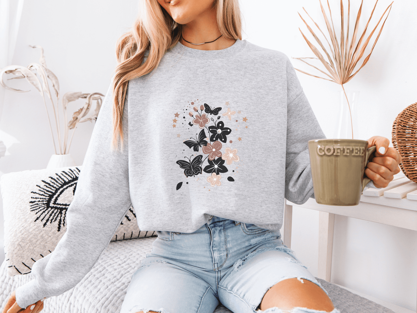 Woman wearing a ash grey sweatshirt featuring elegant black and blush butterflies and flowers, holding a coffee mug – a soft and stylish gift for moms who love whimsical bloom designs.