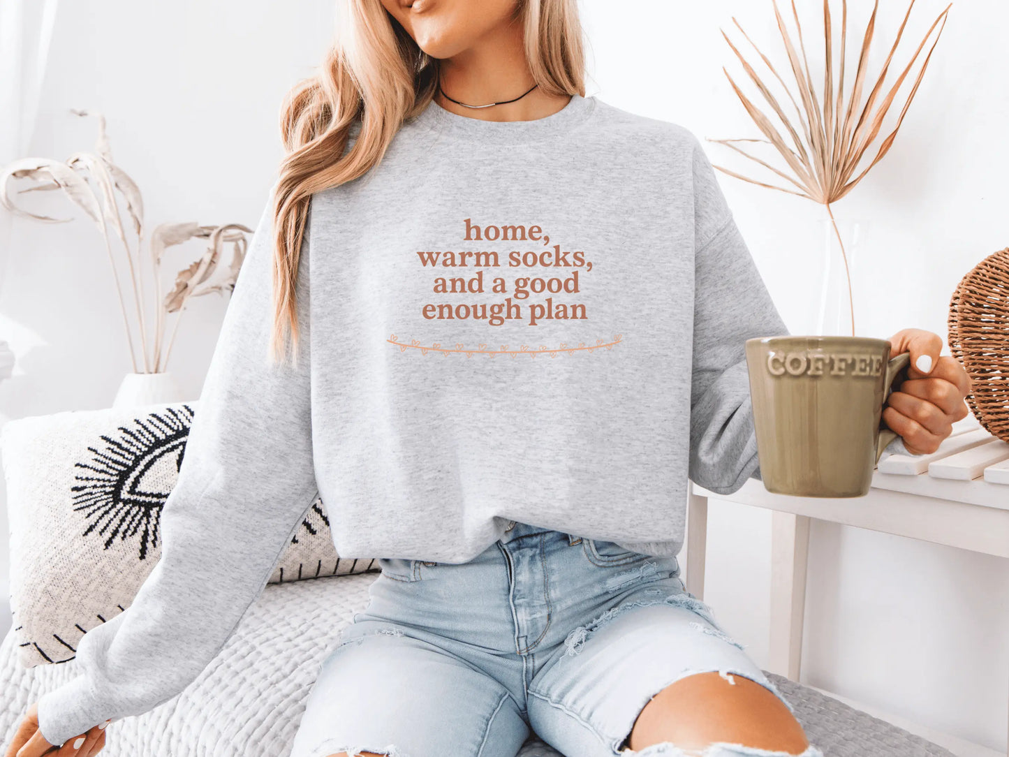 Person wearing a gray sweatshirt with text, holding a coffee mug in a cozy setting.