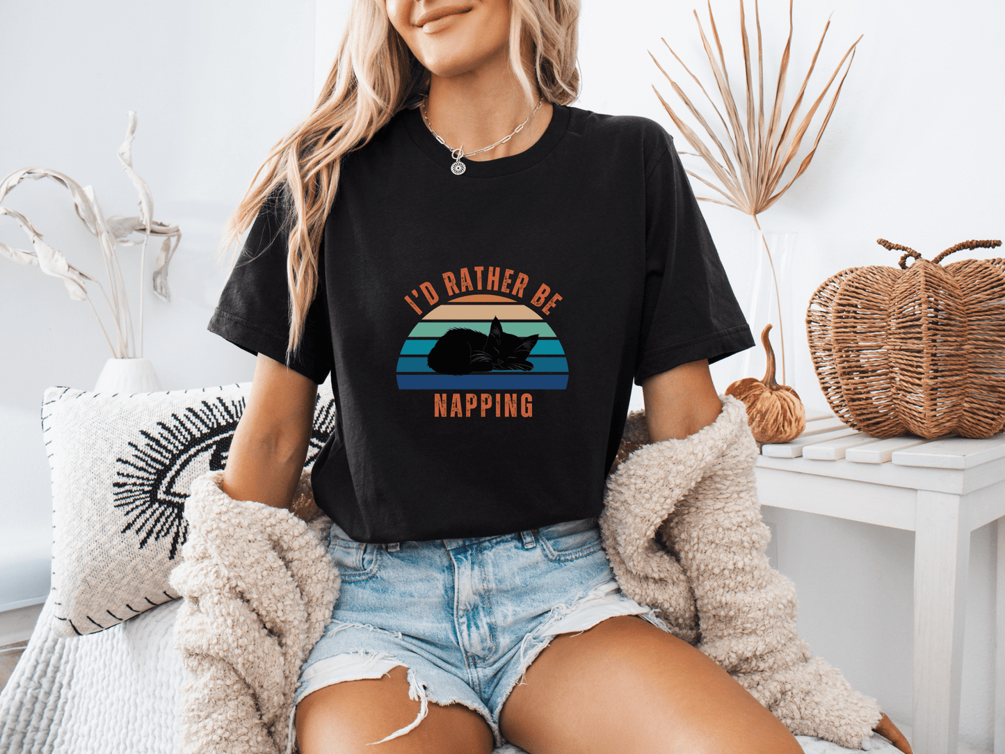 Woman sitting on a couch wearing a black t-shirt with a retro sunset graphic and the phrase "I’d Rather Be Napping," styled with a cozy cardigan and denim shorts.