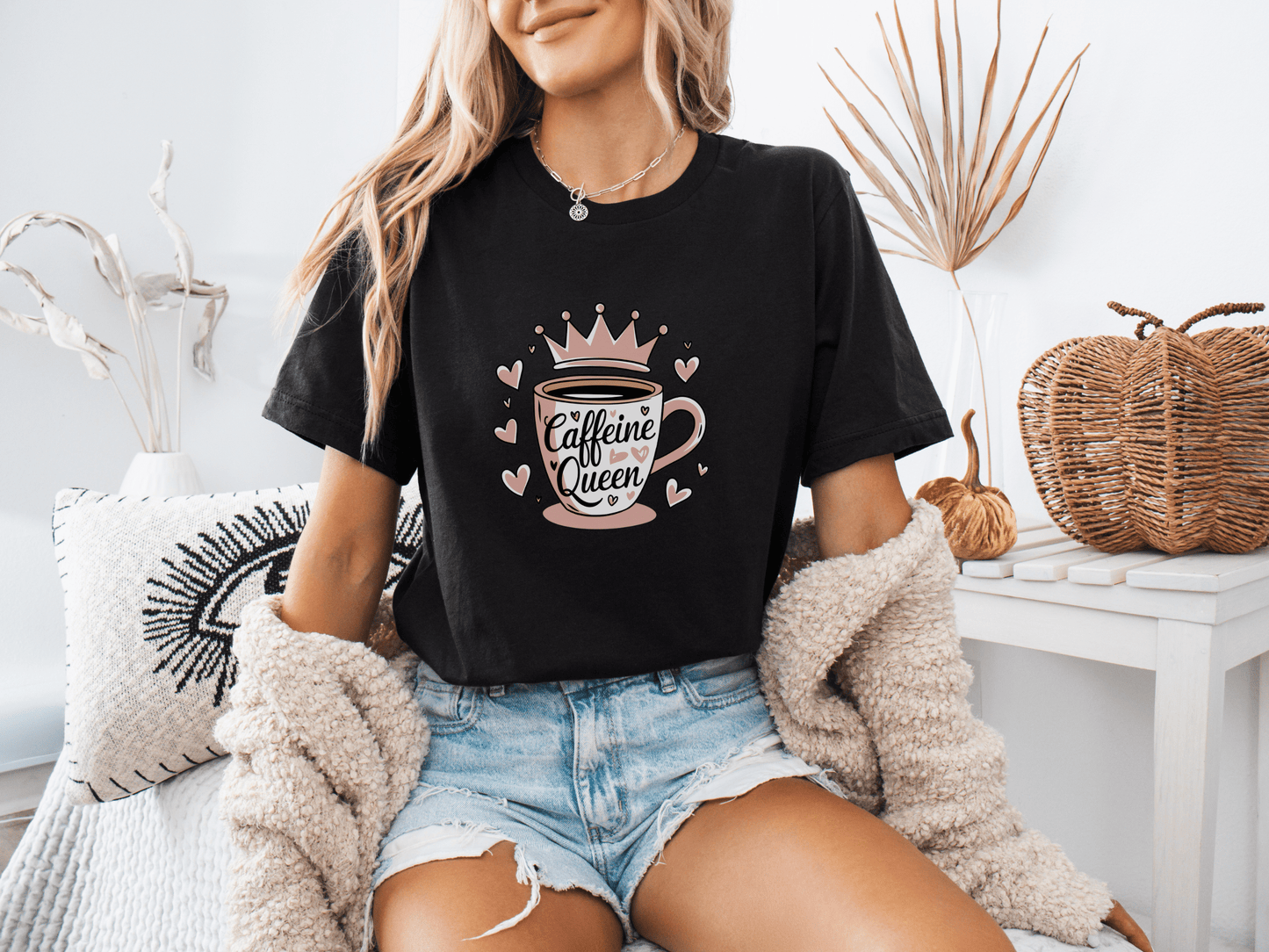 Woman wearing a black “Caffeine Queen” t-shirt with a pink crown coffee cup design, sitting in a cozy room setting