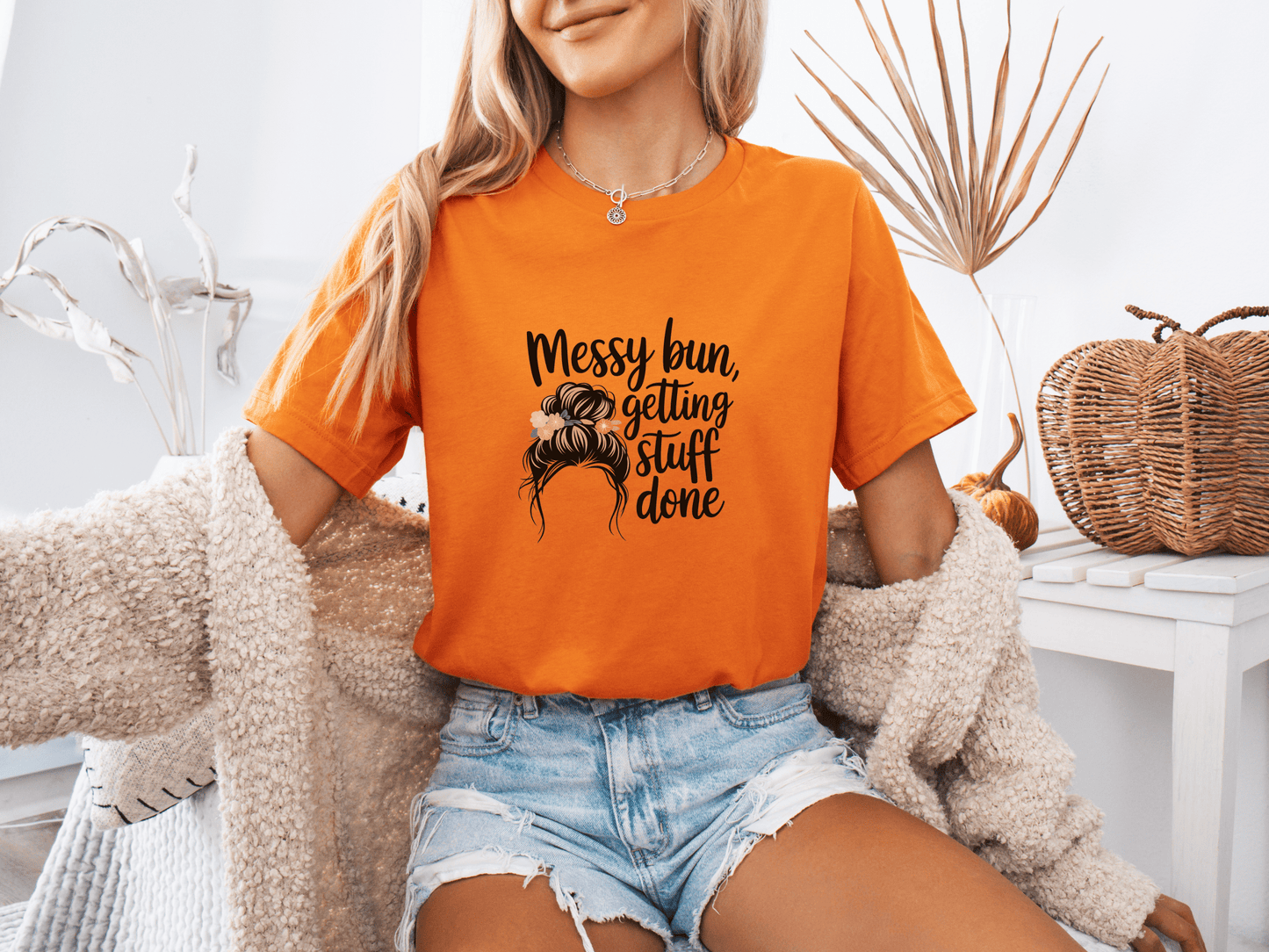 Woman wearing orange “Messy Bun, Getting Stuff Done” t-shirt, posing casually on a bed.