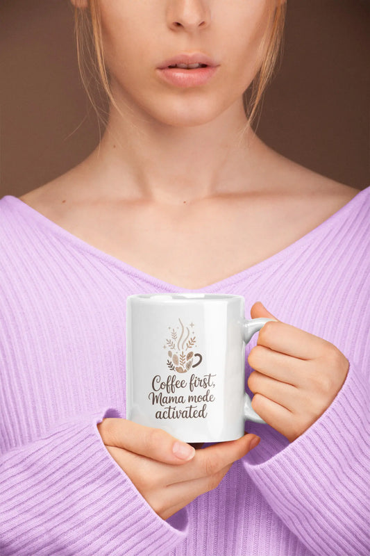 Person holding a mug with 'Coffee first, Mama mode activated' text against a brown background