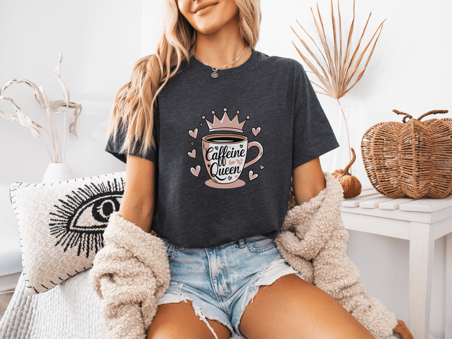 Woman wearing a dark grey heather “Caffeine Queen” t-shirt with a pink crown coffee cup design, sitting in a cozy room setting