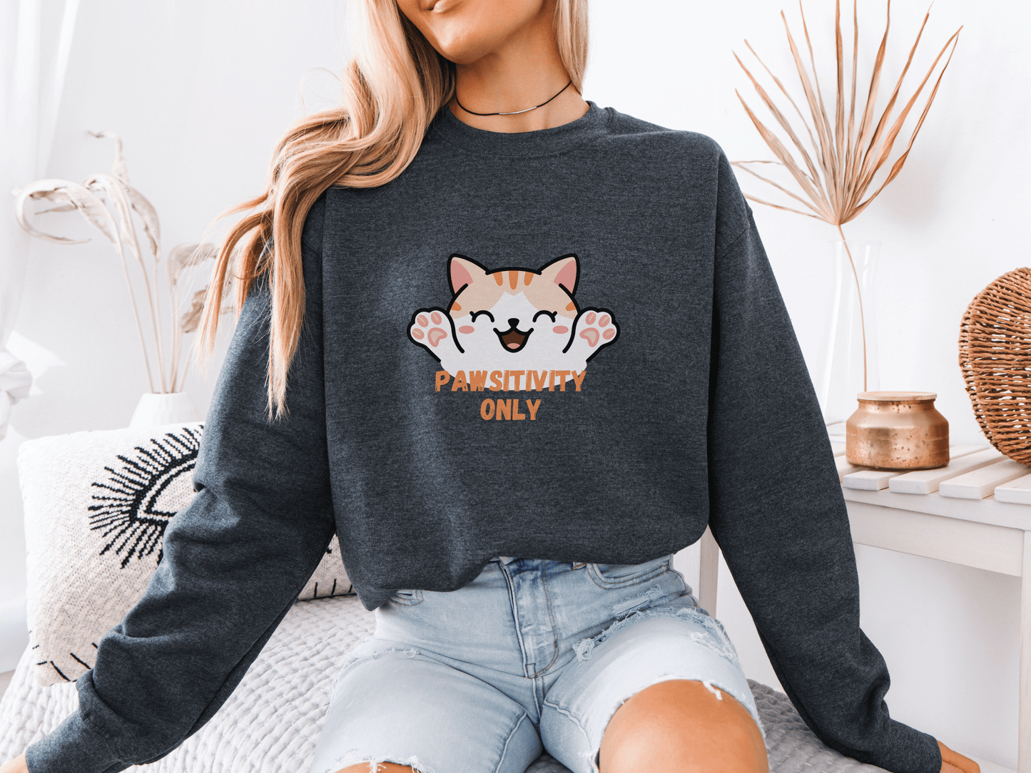 Woman wearing a dark heather sweatshirt with a cheerful orange kitten and the phrase “Pawsitivity Only,” seated in a cozy room