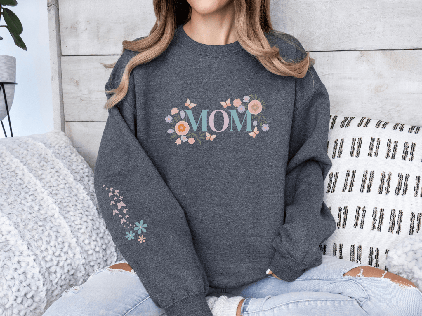 Woman wearing a dark grey sweatshirt with pastel floral “MOM” lettering, seated on a cozy couch with textured pillows