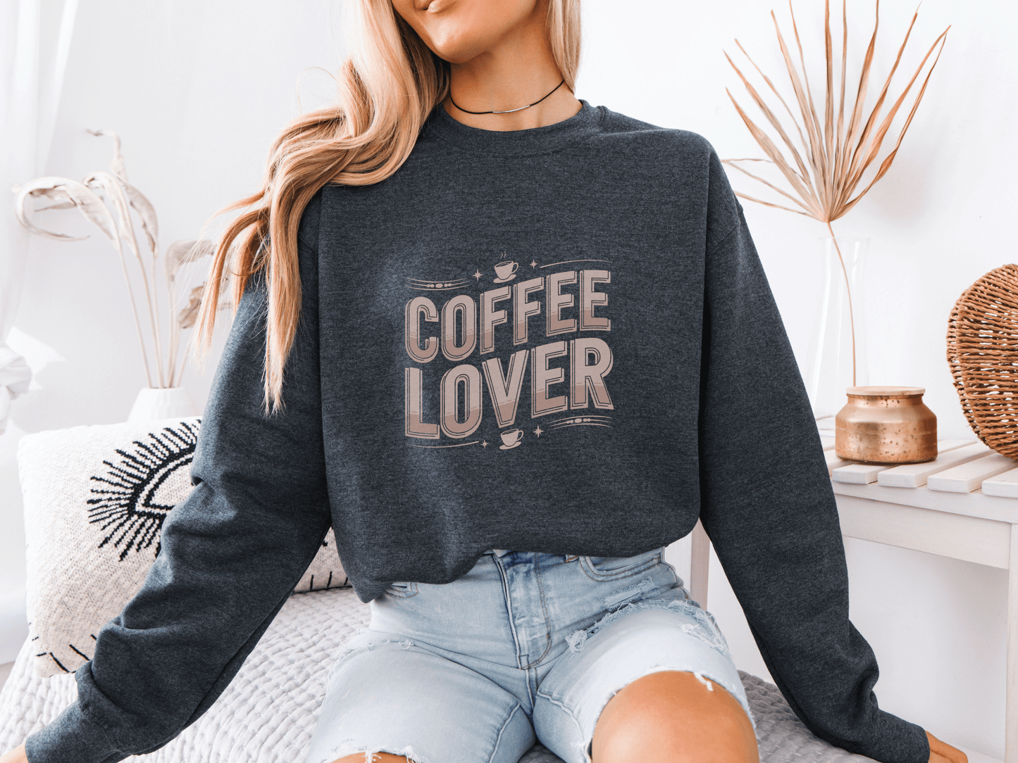 Woman sitting on a bed wearing a dark heather sweatshirt with beige "COFFEE LOVER" text design, styled with ripped light denim shorts in a cozy boho-inspired room with neutral decor and a decorative eye pillow.