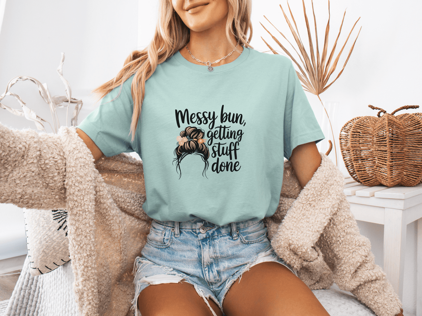 Woman wearing dusty blue “Messy Bun, Getting Stuff Done” t-shirt, posing casually on a bed.