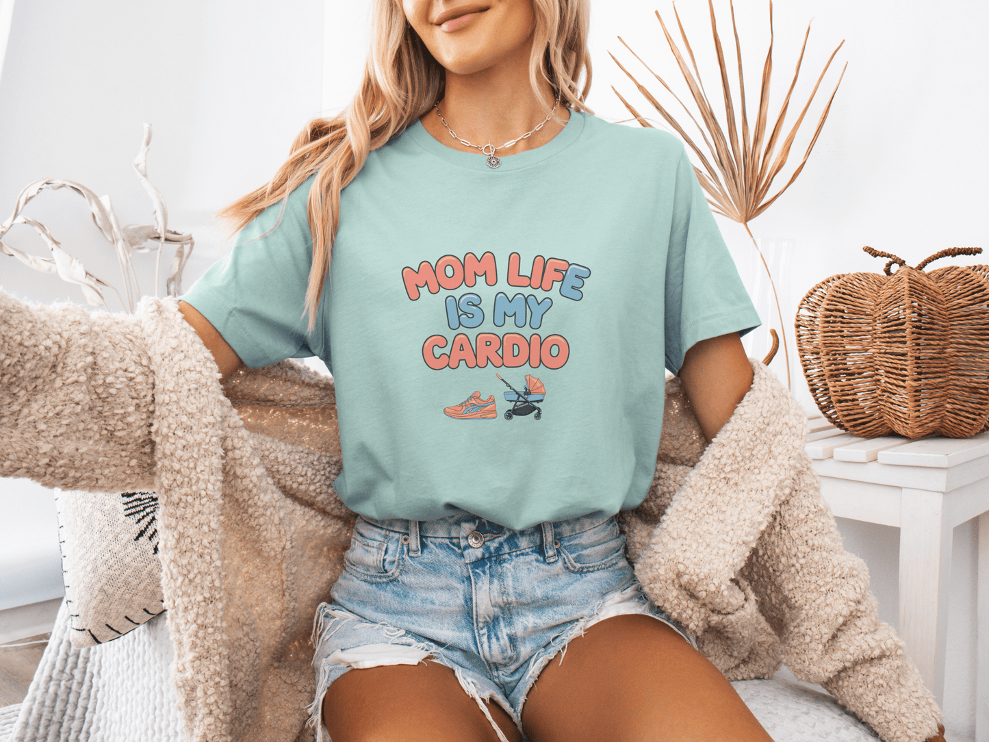Woman wearing a dusty blue graphic tee with the phrase “Mom Life Is My Cardio” in colorful letters, sitting casually in a cozy room