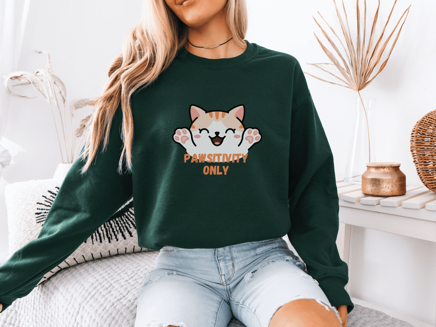Woman wearing a forest green sweatshirt with a cheerful orange kitten and the phrase “Pawsitivity Only,” seated in a cozy room