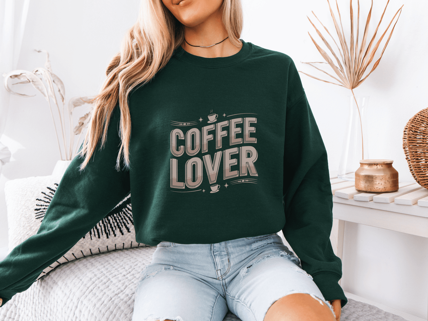Woman sitting on a bed wearing a forest green sweatshirt with beige "COFFEE LOVER" text design, styled with ripped light denim shorts in a cozy boho-inspired room with neutral decor and a decorative eye pillow.