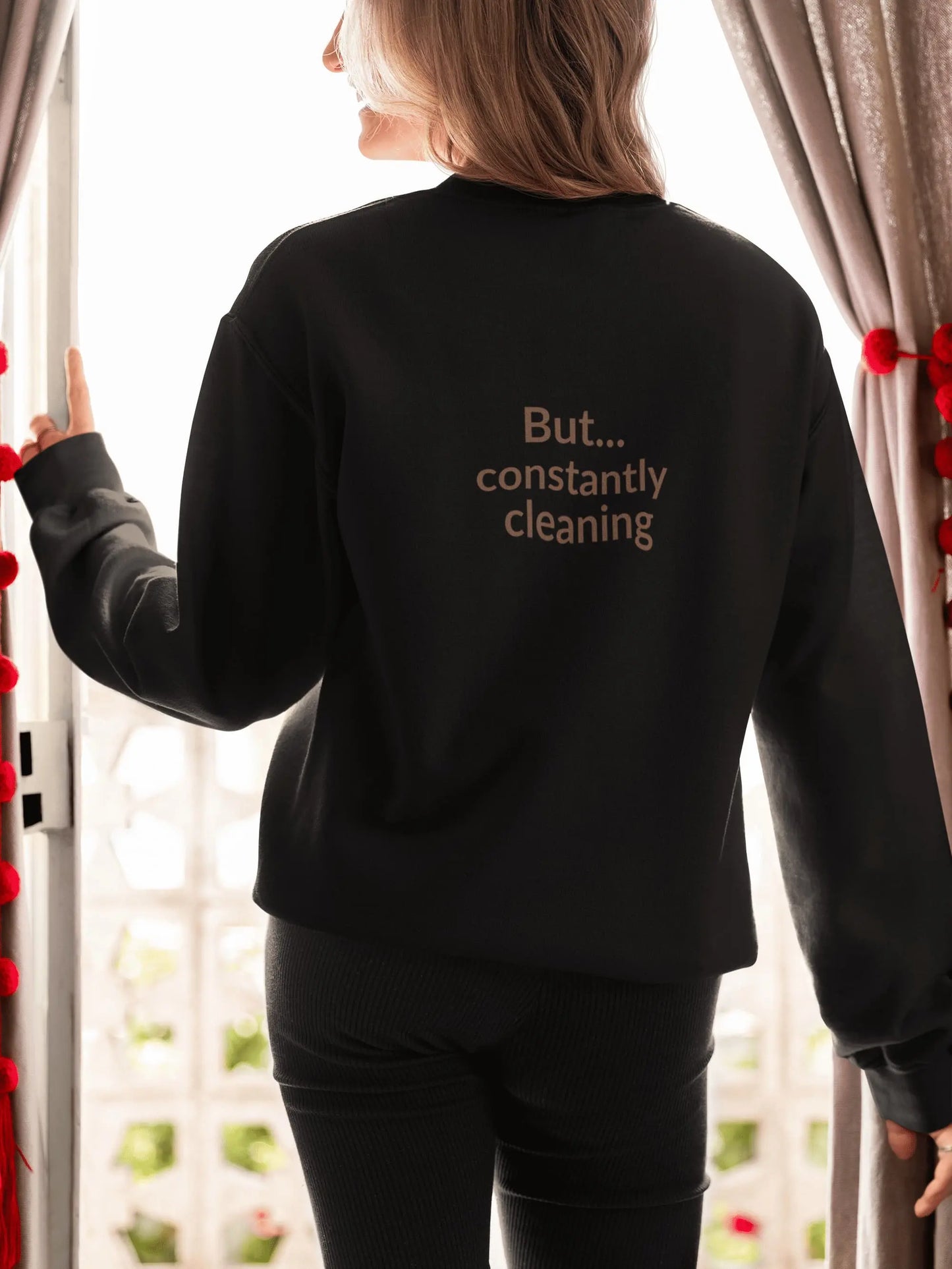 Person wearing a black sweatshirt with text, standing in front of a window with curtains.