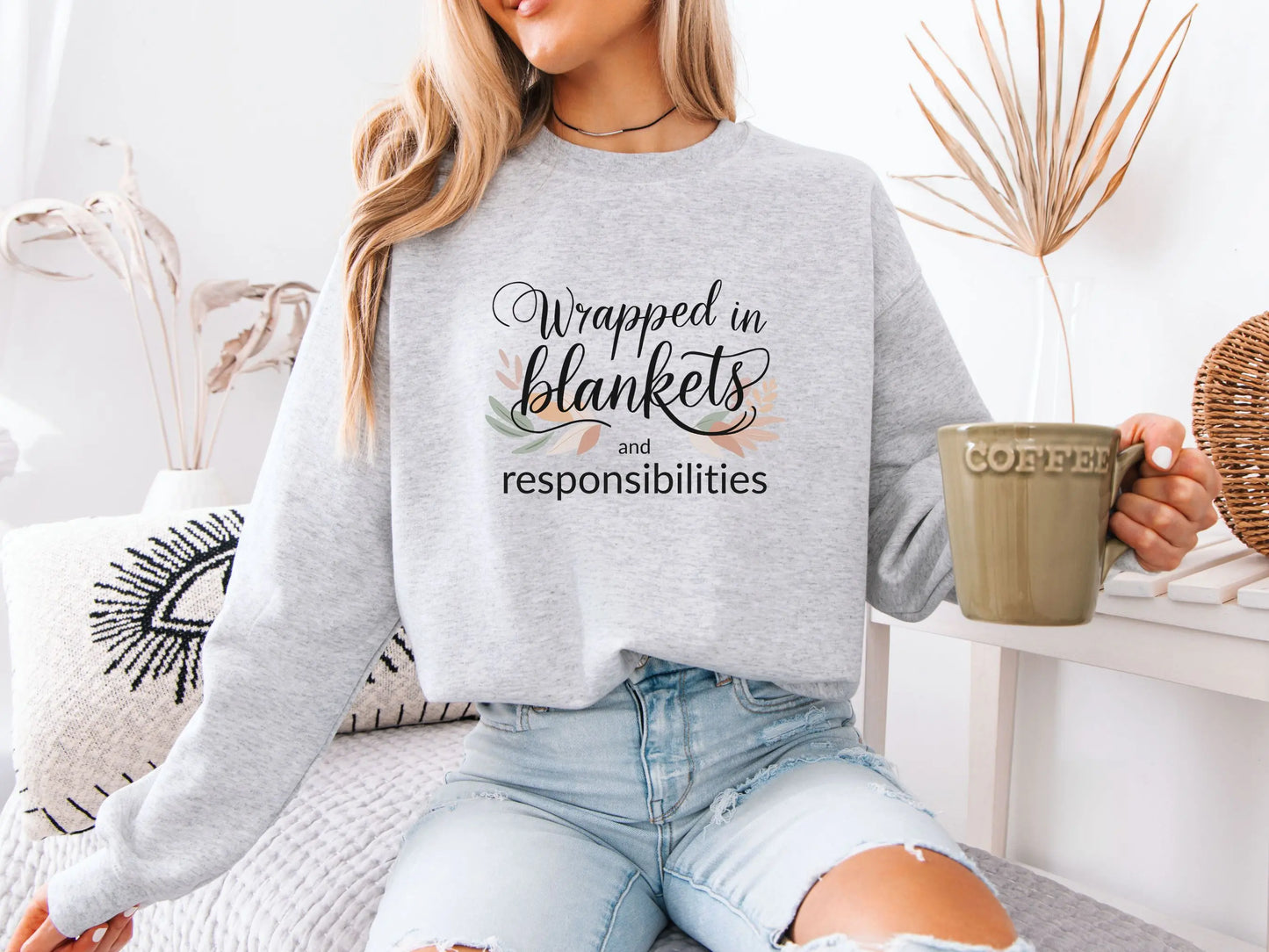 Person wearing a ash grey sweatshirt with text "Wrapped in blankets and responsibilities", sitting on a couch holding a mug.