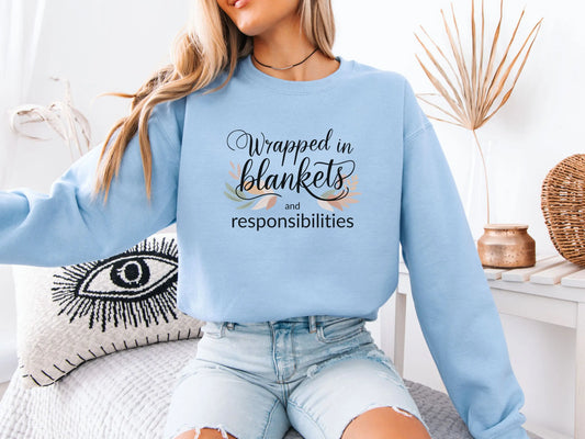 Person wearing a light blue sweatshirt with text "Wrapped in blankets and responsibilities", sitting on a couch holding a mug.