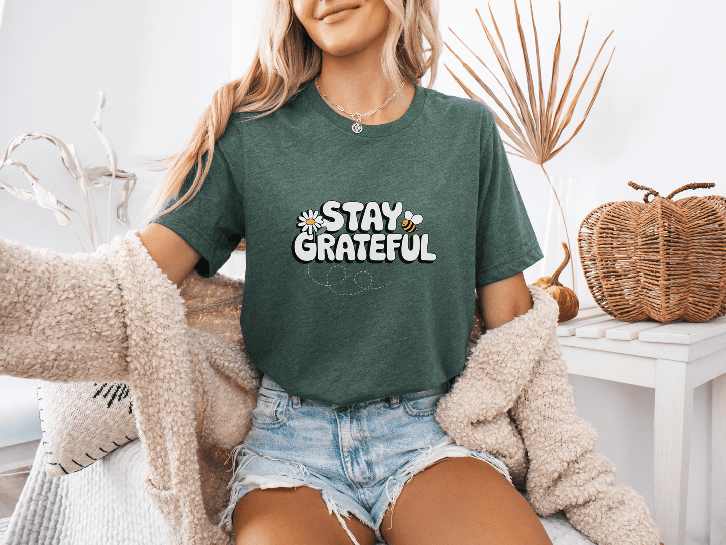 Woman wearing a forest green “Stay Grateful” t-shirt with playful daisy accents, posing in a cozy boho room