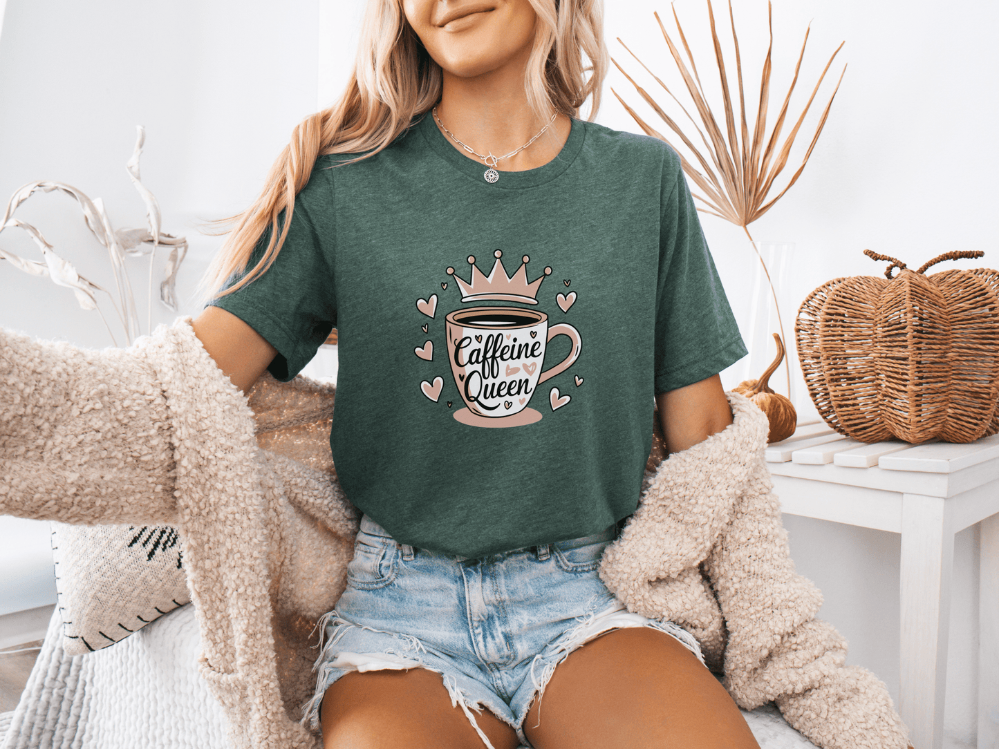 Woman wearing a heather forest green “Caffeine Queen” t-shirt with a pink crown coffee cup design, sitting in a cozy room setting
