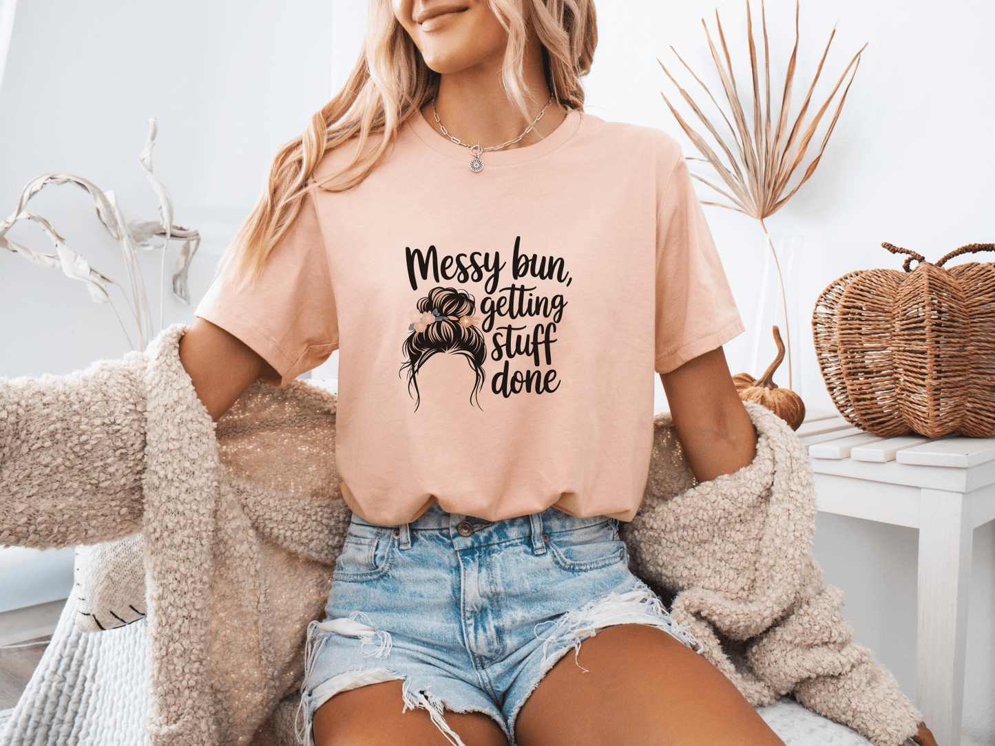 Woman wearing peach “Messy Bun, Getting Stuff Done” t-shirt, posing casually on a bed.