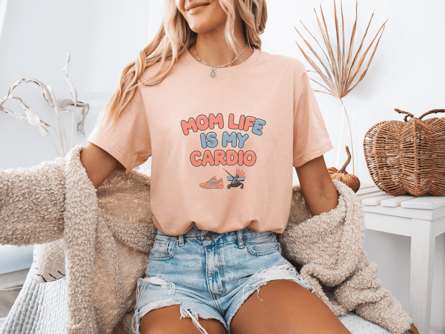 Woman wearing a peach colored graphic tee with the phrase “Mom Life Is My Cardio” in colorful letters, sitting casually in a cozy room