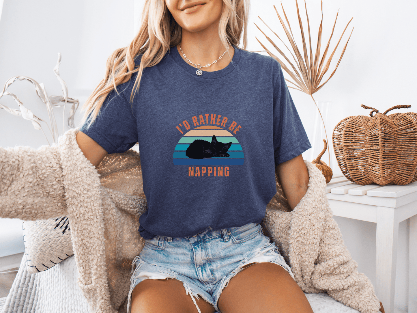 Woman sitting on a couch wearing a heather navy t-shirt with a retro sunset graphic and the phrase "I’d Rather Be Napping," styled with a cozy cardigan and denim shorts.