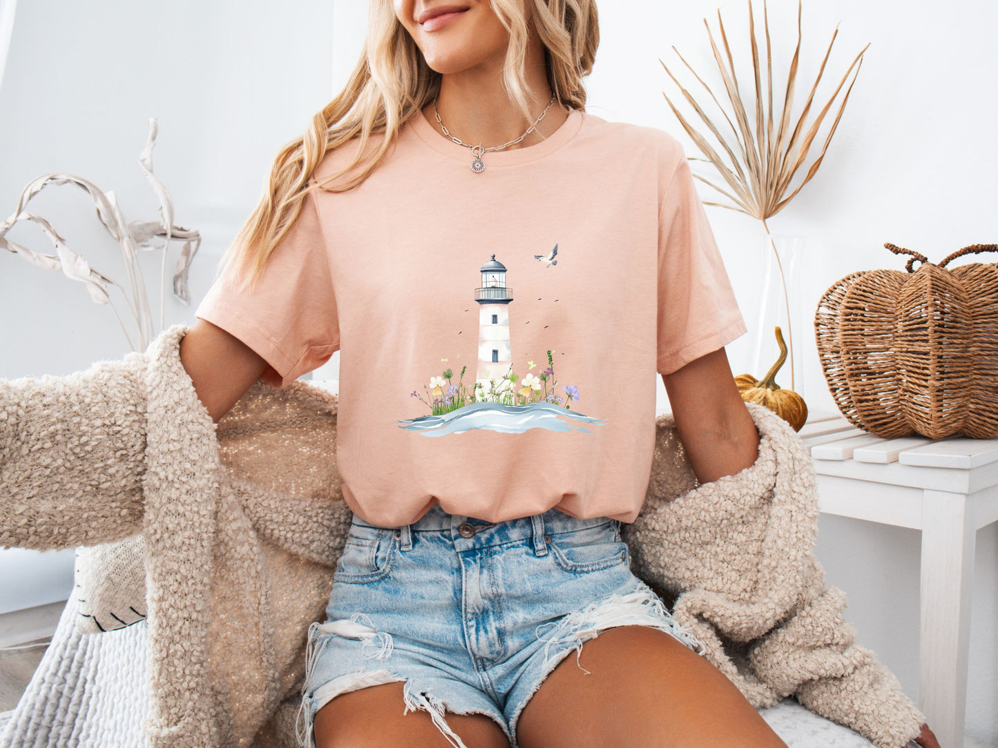 Woman sitting on a couch wearing a heather peach t-shirt with a whimsical lighthouse and floral design, paired with denim shorts and a cozy knit cardigan, in a calm and stylish home setting.