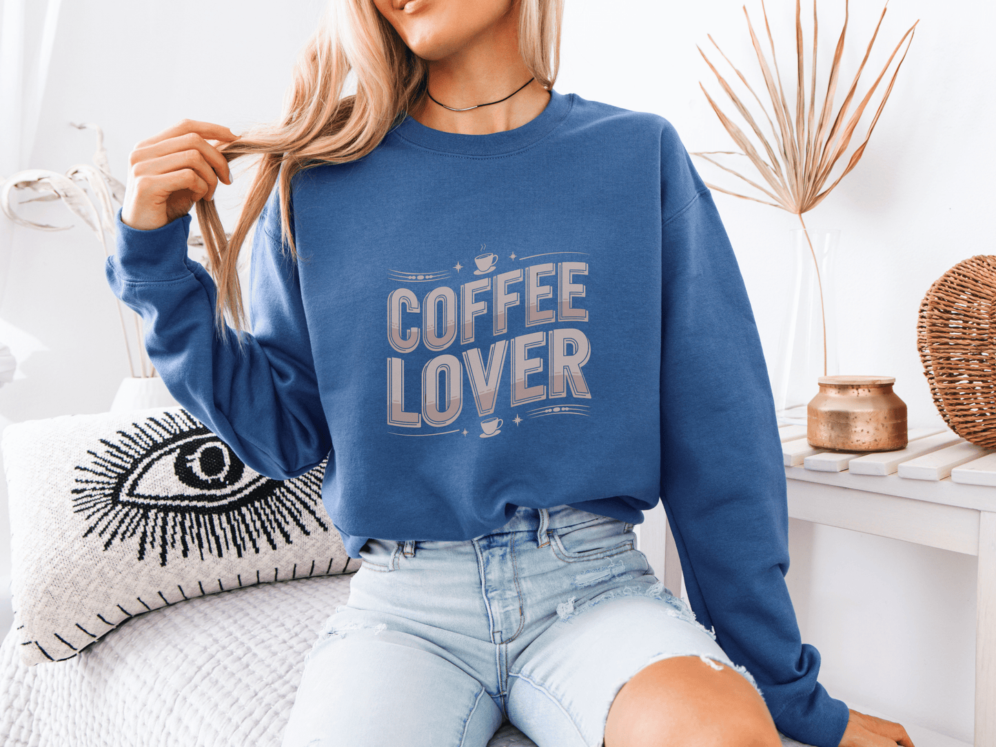 Woman sitting on a bed wearing a indigo blue sweatshirt with beige "COFFEE LOVER" text design, styled with ripped light denim shorts in a cozy boho-inspired room with neutral decor and a decorative eye pillow.