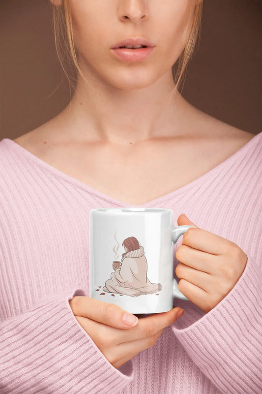 Person holding a mug with a design of a person wrapped in a blanket holding a cup, wearing a pink sweater.