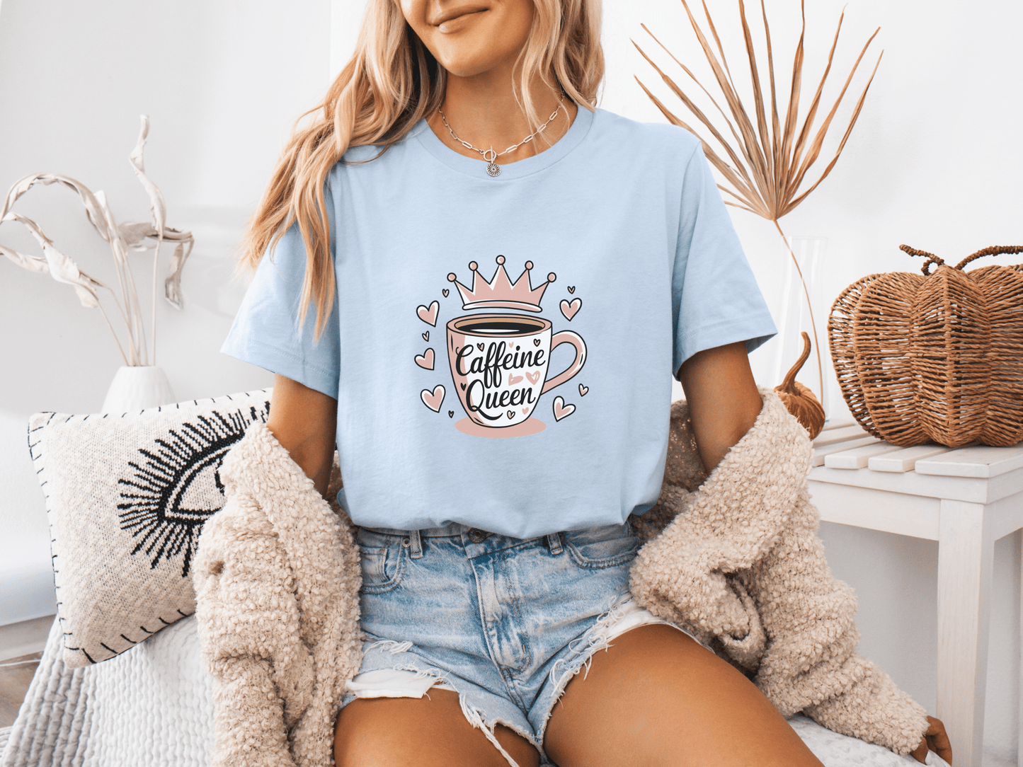 Woman wearing a light blue “Caffeine Queen” t-shirt with a pink crown coffee cup design, sitting in a cozy room setting