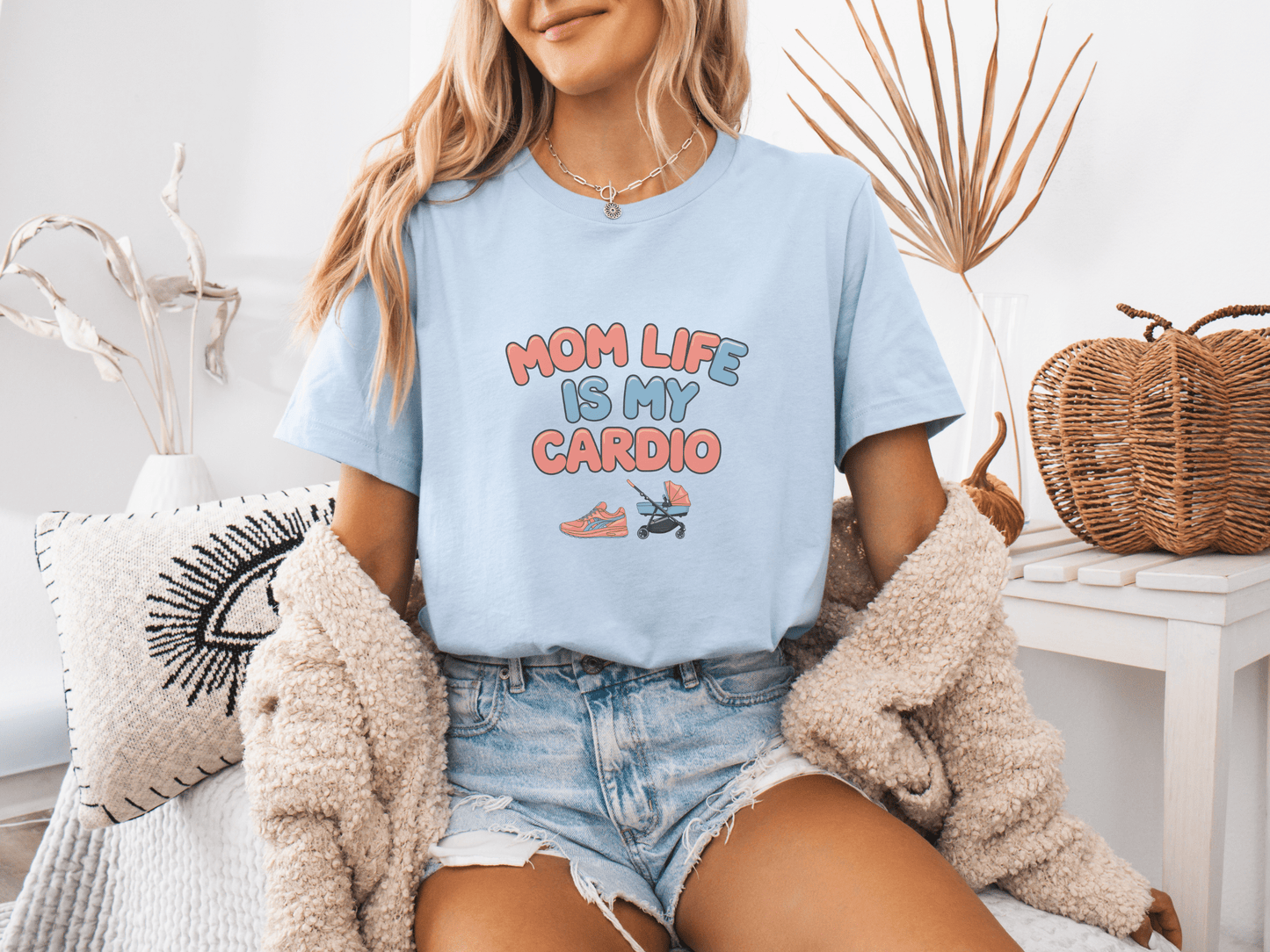 Woman wearing a light blue graphic tee with the phrase “Mom Life Is My Cardio” in colorful letters, sitting casually in a cozy room