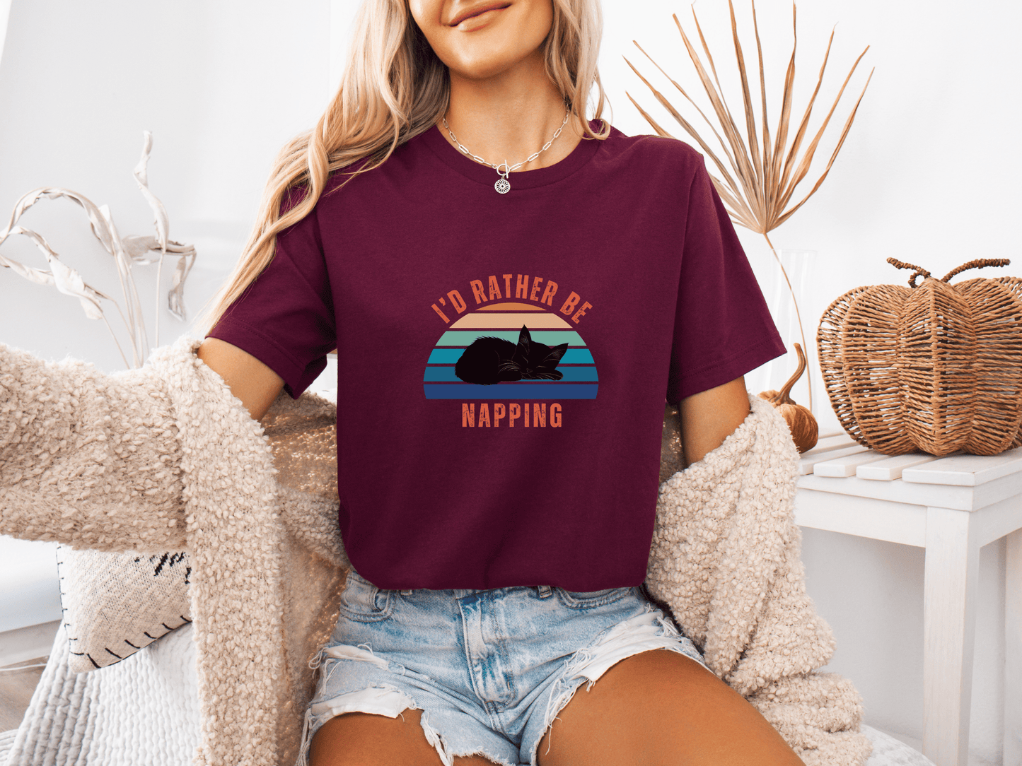 Woman sitting on a couch wearing a maroon color t-shirt with a retro sunset graphic and the phrase "I’d Rather Be Napping," styled with a cozy cardigan and denim shorts.