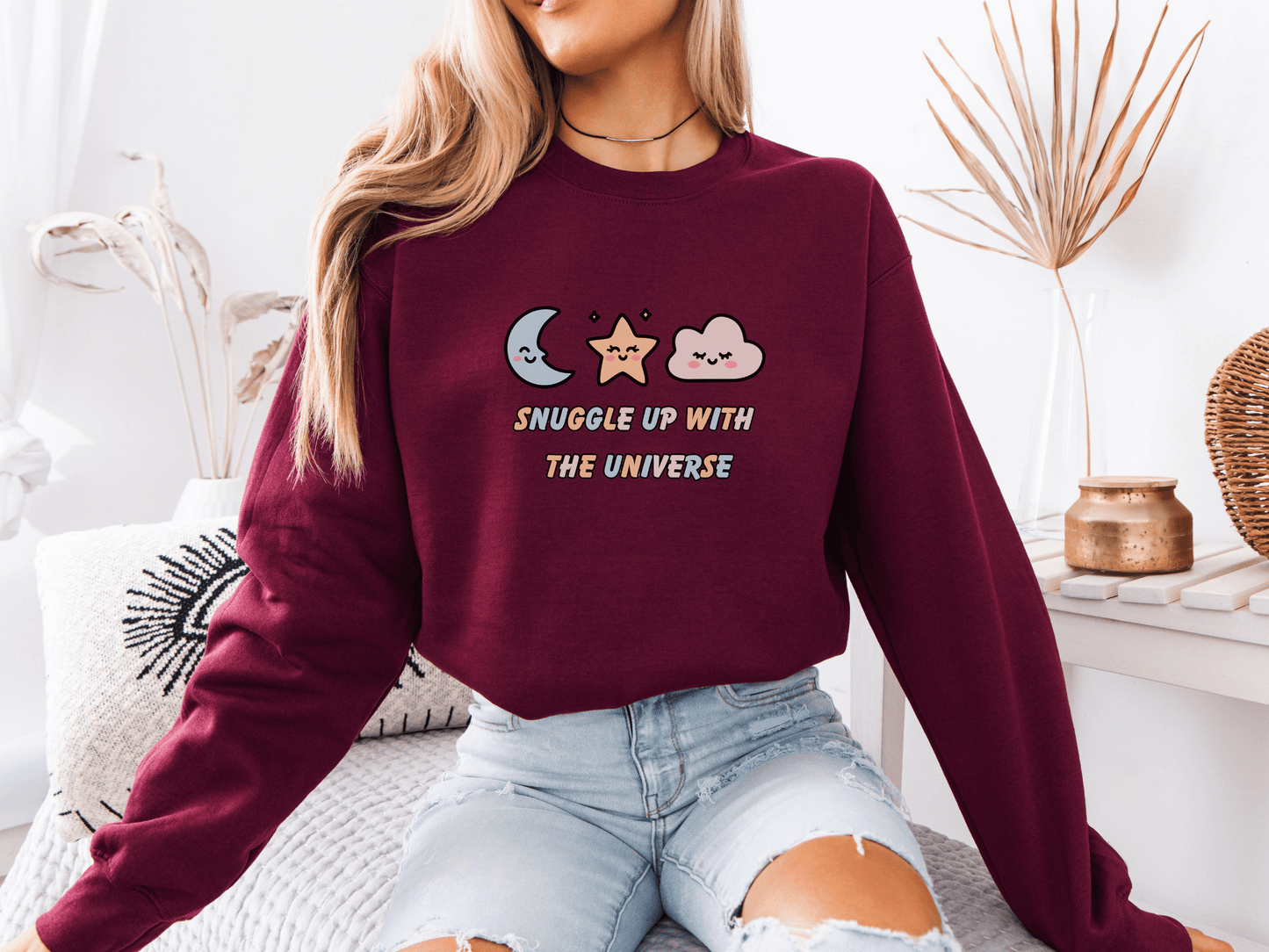 Woman wearing a maroon “Snuggle Up With the Universe” sweatshirt, holding a coffee mug and relaxing on a cozy bed