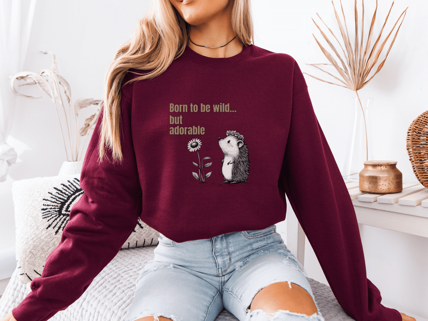 Woman wearing a maroon red sweatshirt with “Born to be wild… but adorable” text and a cute hedgehog graphic, sitting on a cozy white couch.