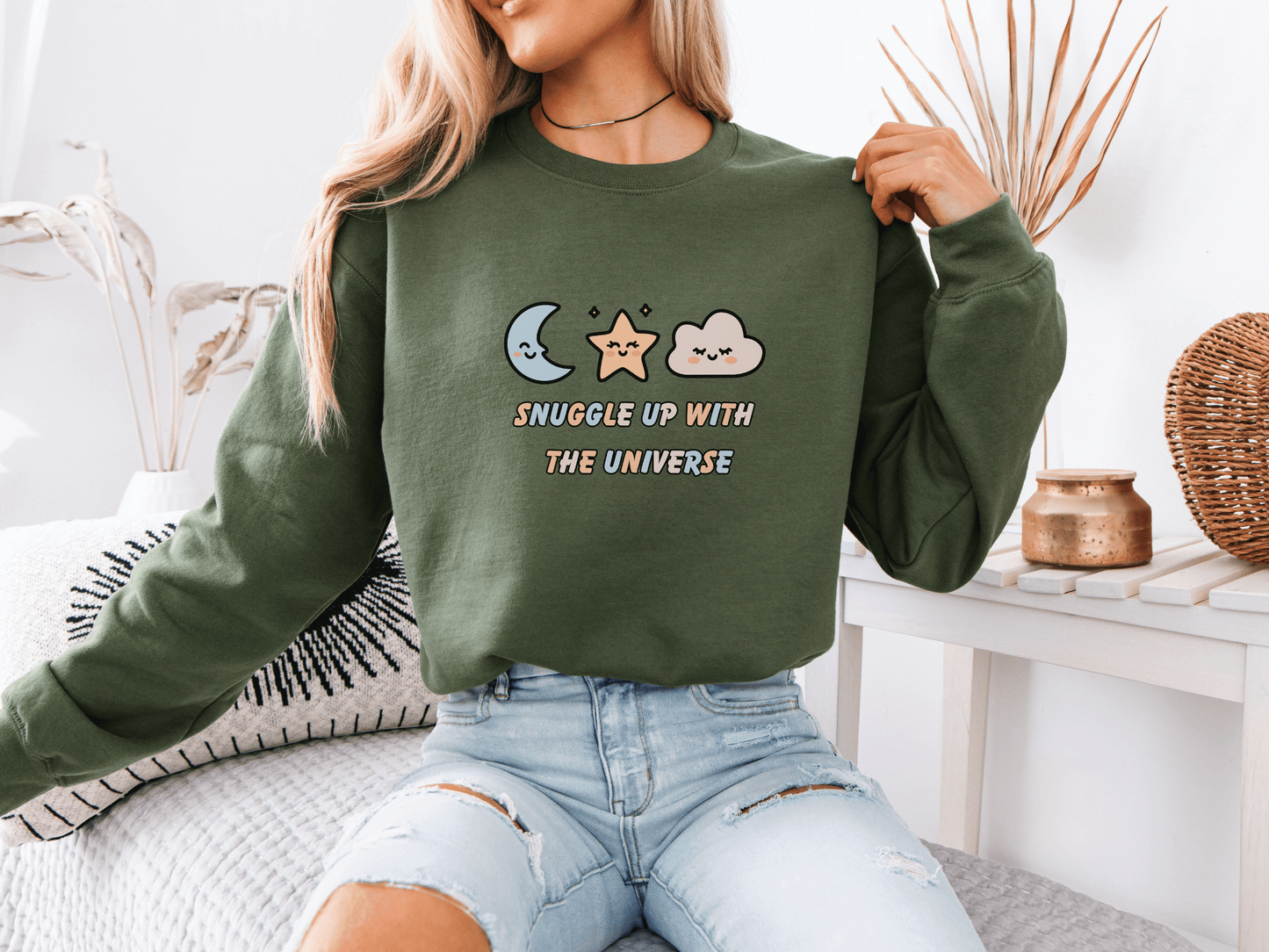 Woman wearing a military green “Snuggle Up With the Universe” sweatshirt, holding a coffee mug and relaxing on a cozy bed