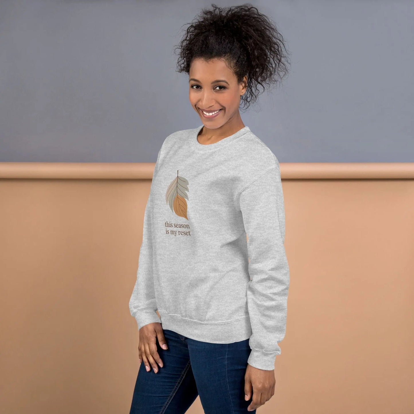 Woman wearing a gray sweatshirt with a leaf design and text "this season is my reset" on a plain background