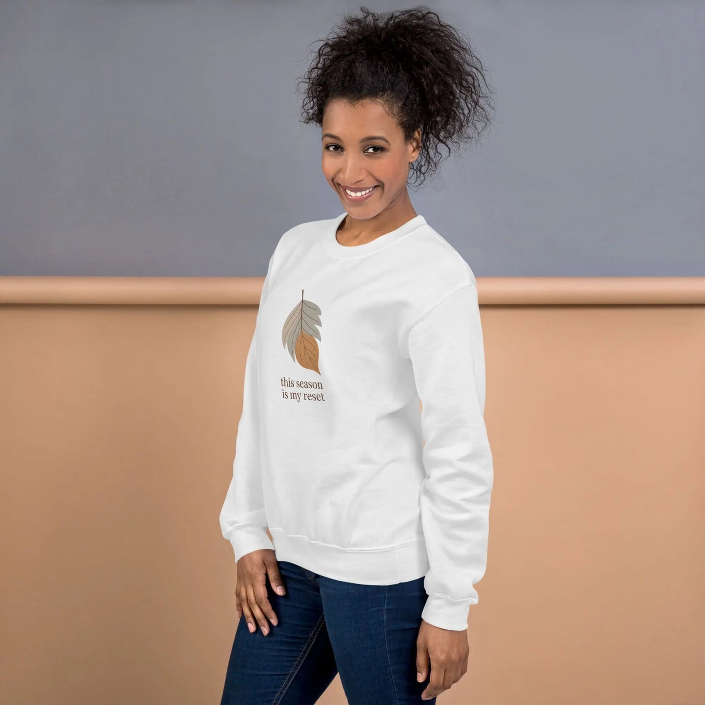 Woman wearing a white sweatshirt with a leaf design and text "this season is my reset" on a plain background