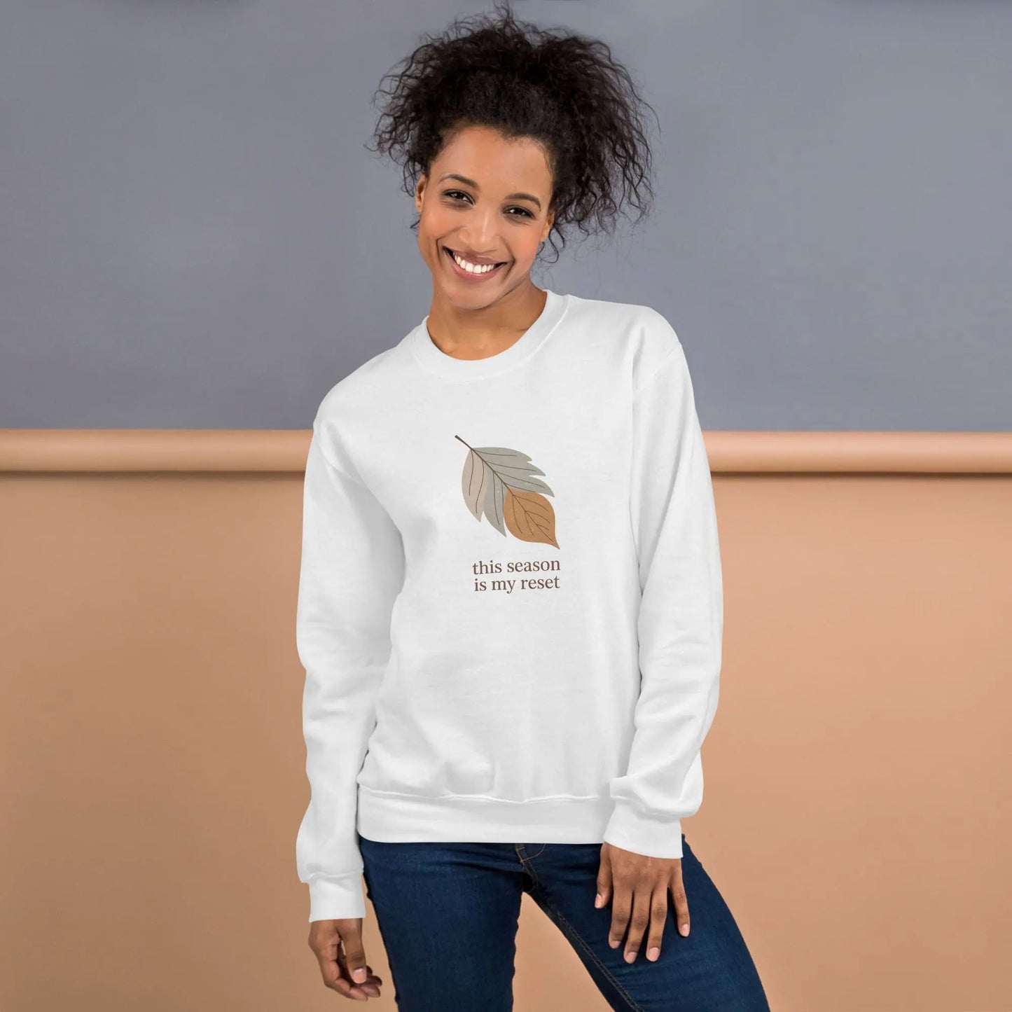 Woman wearing a white sweatshirt with a leaf design and text "this season is my reset" standing against a neutral background