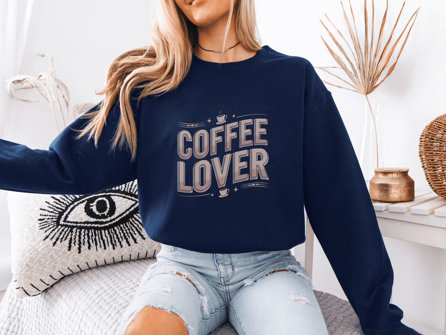 Woman sitting on a bed wearing a navy blue sweatshirt with beige "COFFEE LOVER" text design, styled with ripped light denim shorts in a cozy boho-inspired room with neutral decor and a decorative eye pillow.