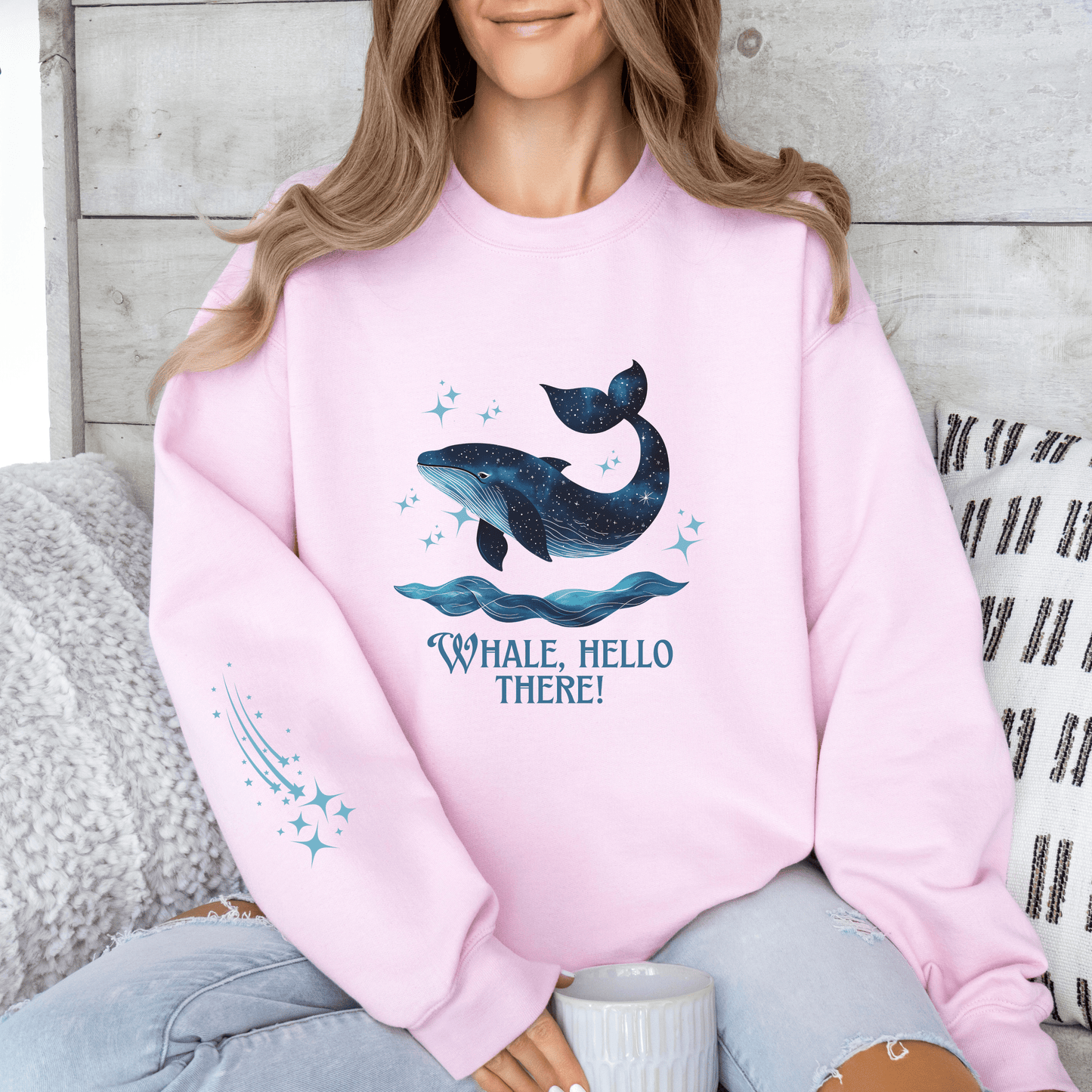 Woman wearing a soft pink sweatshirt featuring a galaxy-style whale jumping from ocean waves with the quote “Whale, Hello There!”