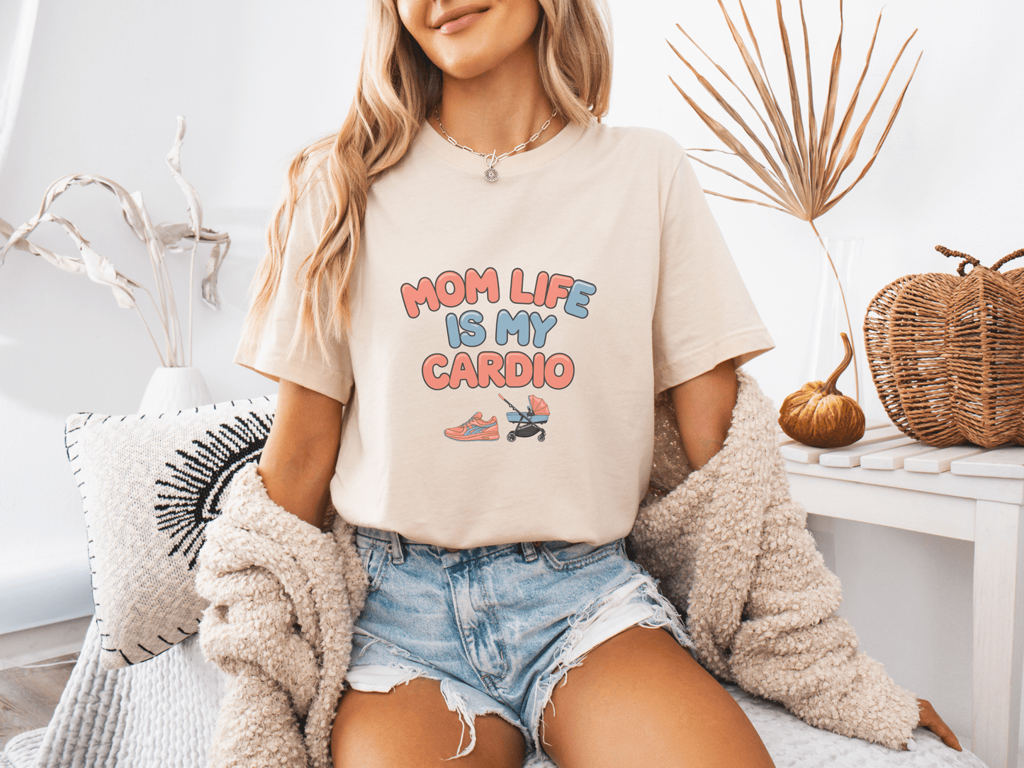 Woman wearing a soft cream graphic tee with the phrase “Mom Life Is My Cardio” in colorful letters, sitting casually in a cozy room