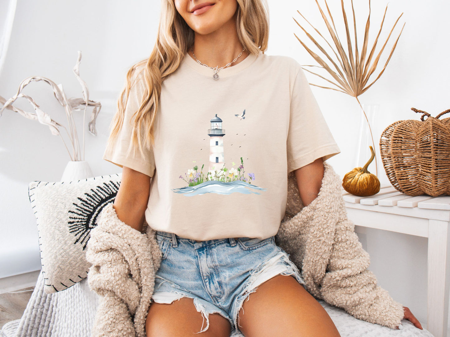 Woman sitting on a couch wearing a soft cream t-shirt with a whimsical lighthouse and floral design, paired with denim shorts and a cozy knit cardigan, in a calm and stylish home setting.
