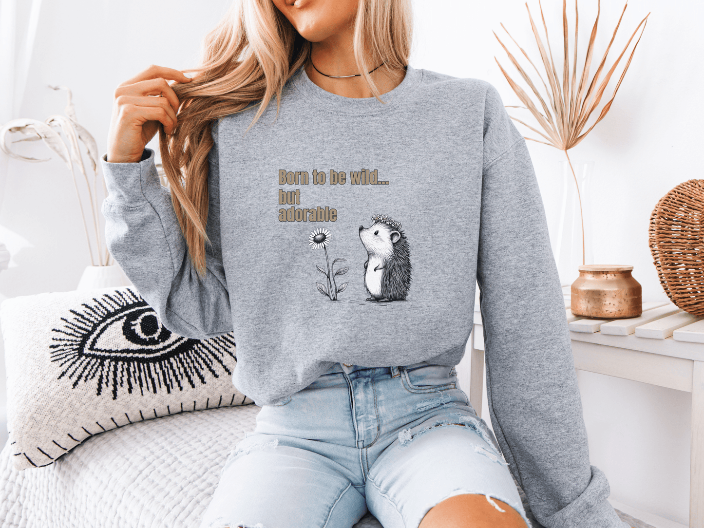 Woman wearing a light gray sweatshirt with “Born to be wild… but adorable” text and a cute hedgehog graphic, sitting on a cozy white couch.