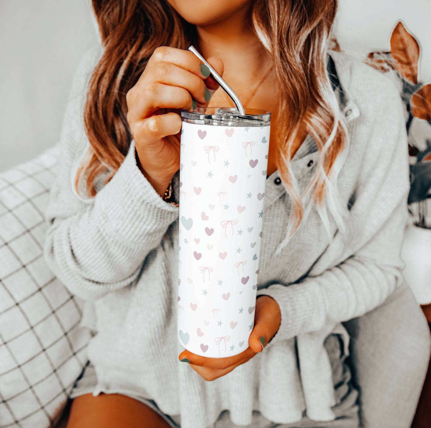 Woman sitting on a couch holding a tall stainless steel tumbler with pastel heart, star, and bow pattern; wearing a cozy gray sweater with long wavy hair partially visible.