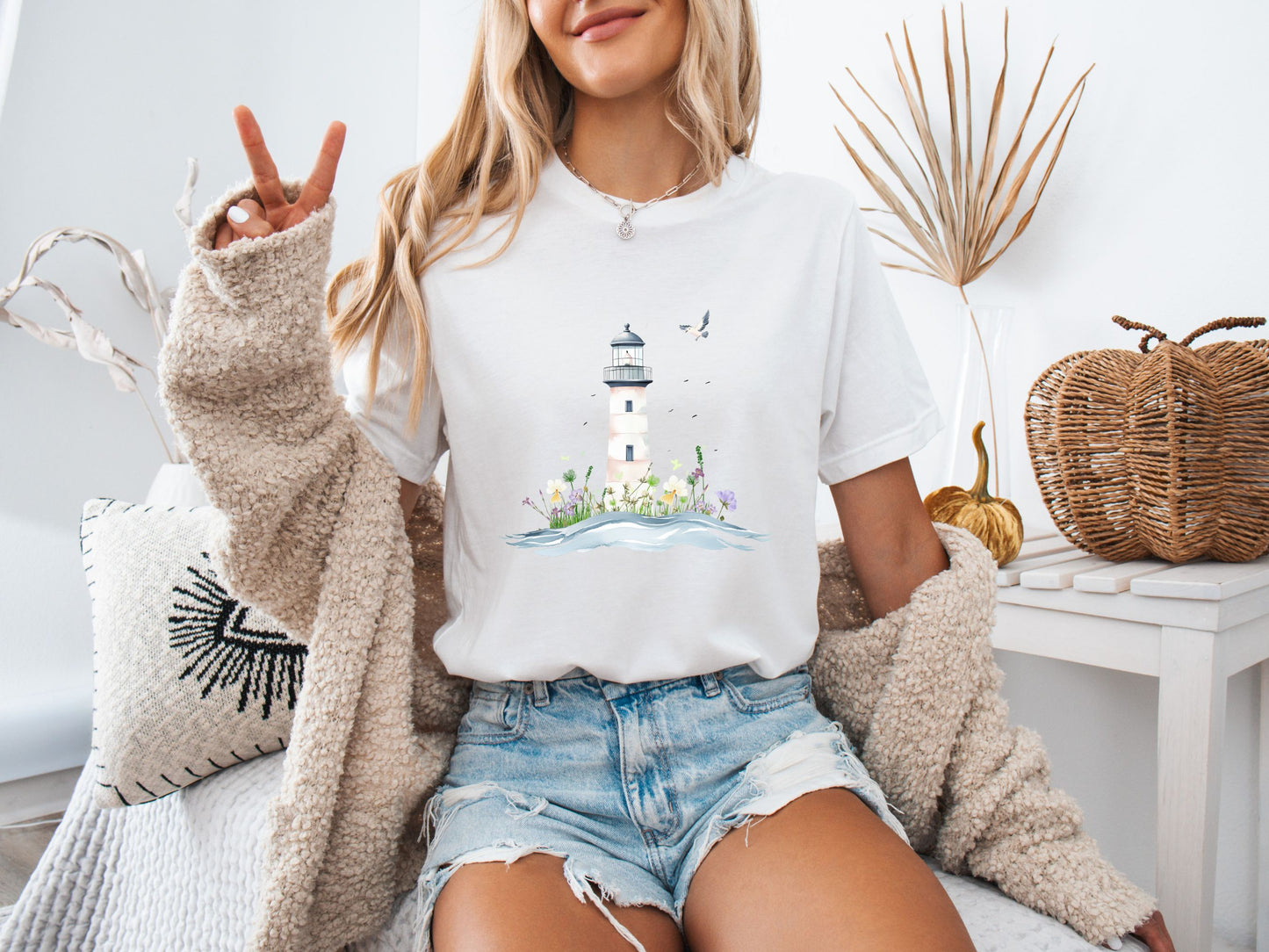 Woman sitting on a couch wearing a white t-shirt with a whimsical lighthouse and floral design, paired with denim shorts and a cozy knit cardigan, in a calm and stylish home setting.