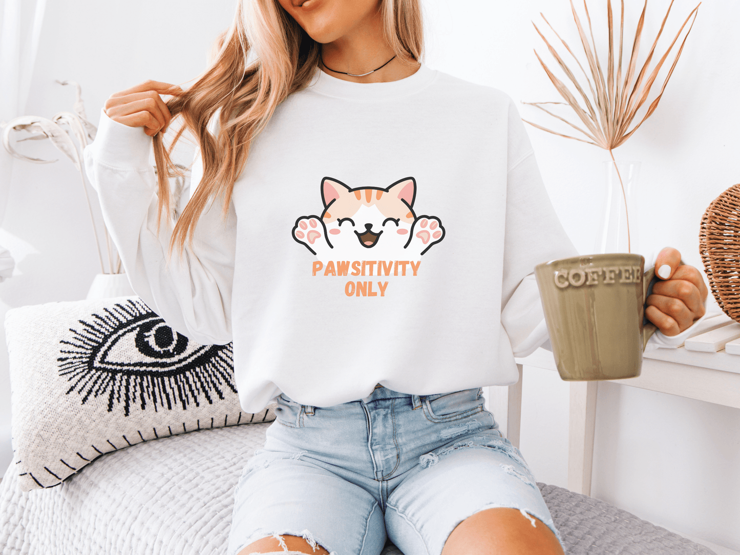 Woman wearing a white sweatshirt with a smiling cat and the phrase “Pawsitivity Only,” holding a green coffee mug in a cozy boho living space