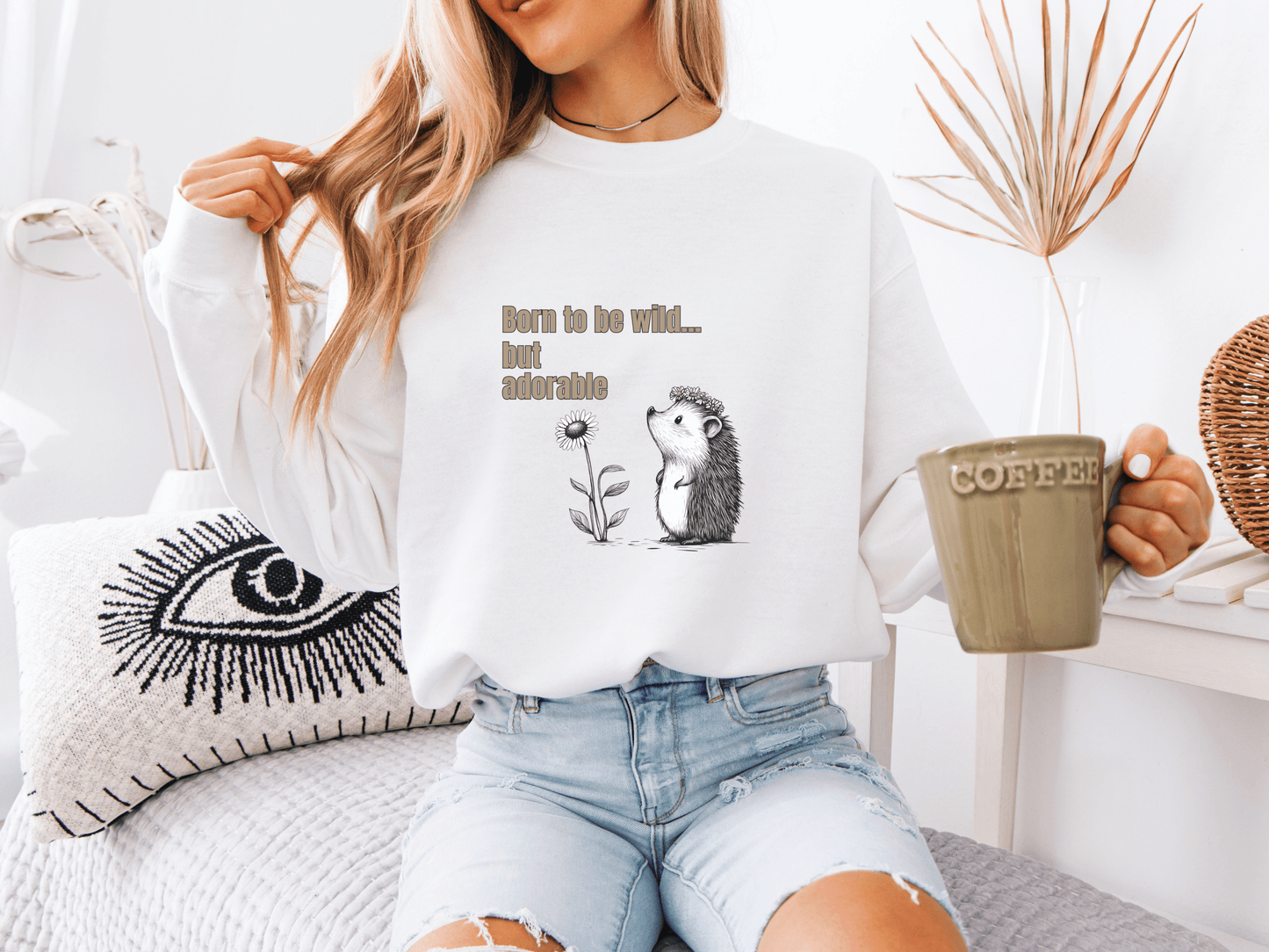 Woman wearing a white sweatshirt with “Born to be wild… but adorable” text and a cute hedgehog graphic, sitting on a cozy white couch.