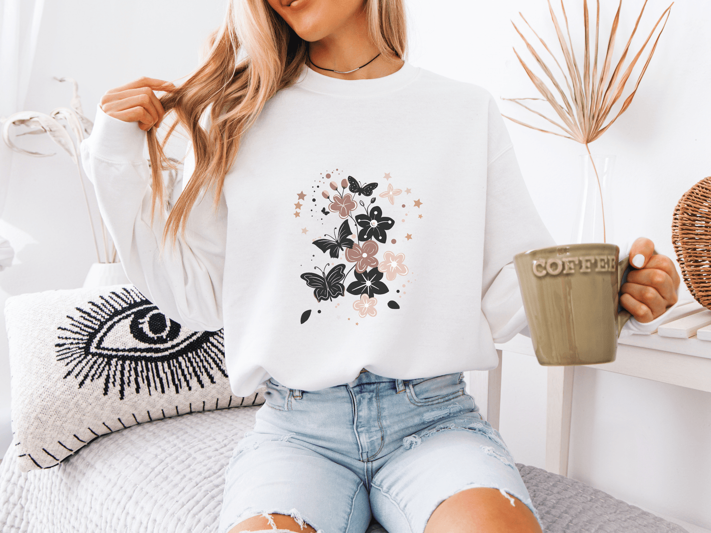 Woman wearing a white sweatshirt featuring elegant black and blush butterflies and flowers, holding a coffee mug – a soft and stylish gift for moms who love whimsical bloom designs.