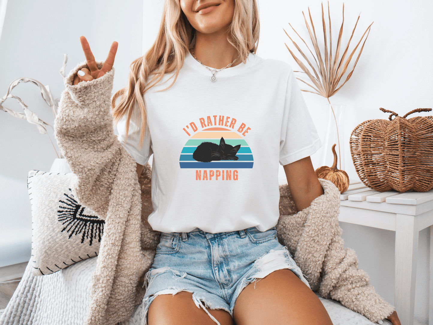 Woman sitting on a couch wearing a white t-shirt with a retro sunset graphic and the phrase "I’d Rather Be Napping," styled with a cozy cardigan and denim shorts.