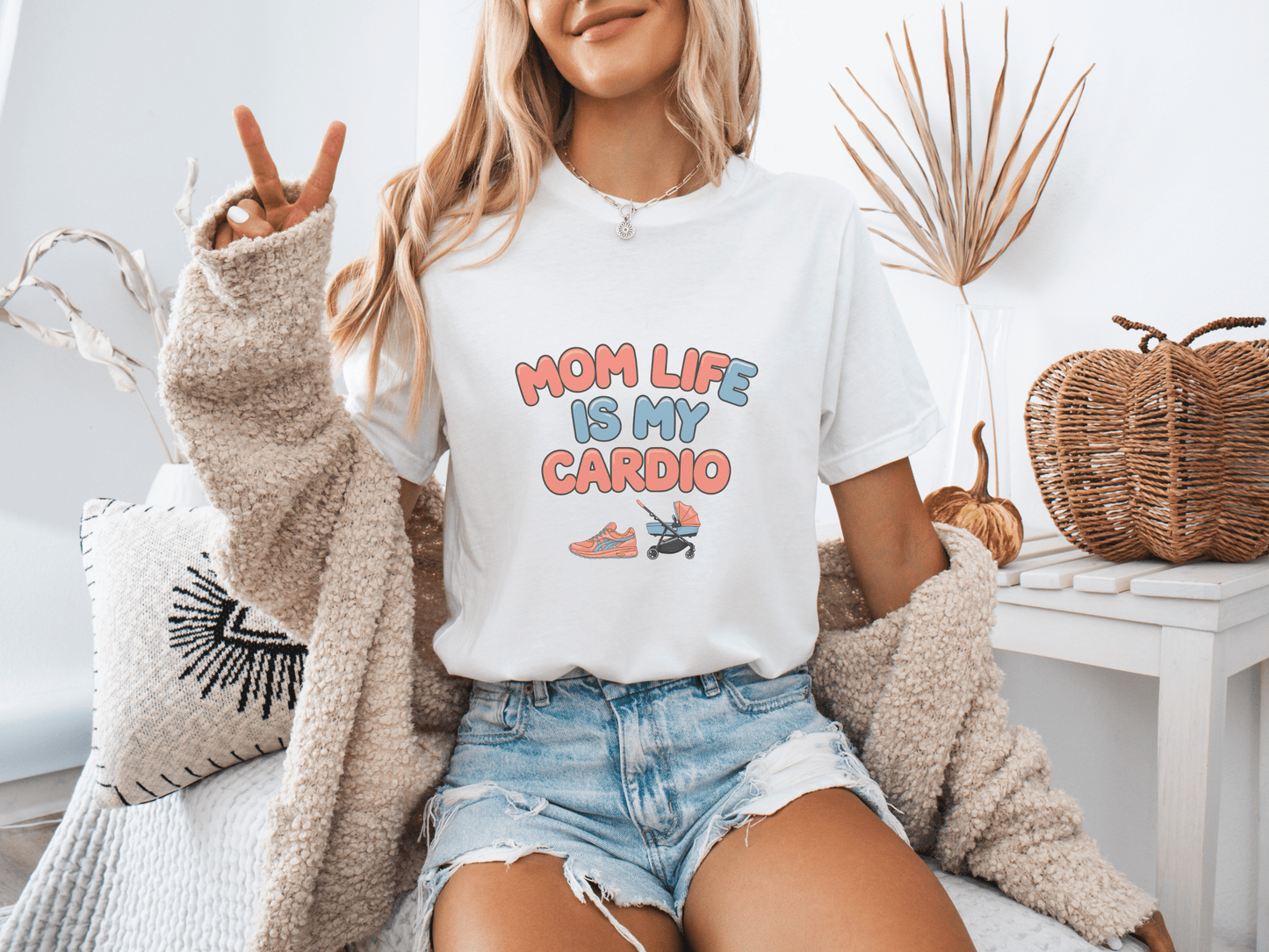 Woman wearing a white graphic tee with the phrase “Mom Life Is My Cardio” in colorful letters, sitting casually in a cozy room